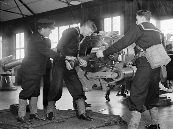 New recruits to the Navy carrying out their training at HMS Raleigh during the wartime period pictured loading a projectile into the breech of a 6 inch gun. The gun is indoors in a large room. These photographs are part of a series documenting the progress of a young sailor, John Smith, through his basic training. Source Imperial War Museum collection