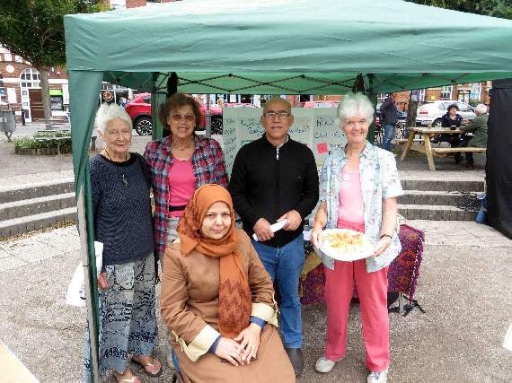 Explaining about the Refugees in Crediton organisation, 25 years-old this year, were (left to right) Pat Barker, Caroline Romijn, Jenny Berg holding a tray of cakes and two Syrian Kurdish refugees. SR 1955