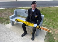 <p>Bob Graham, with the Royal Hampshire Regimental Standard, tries out the granite bench presented to the village of Asnelles in France</p>