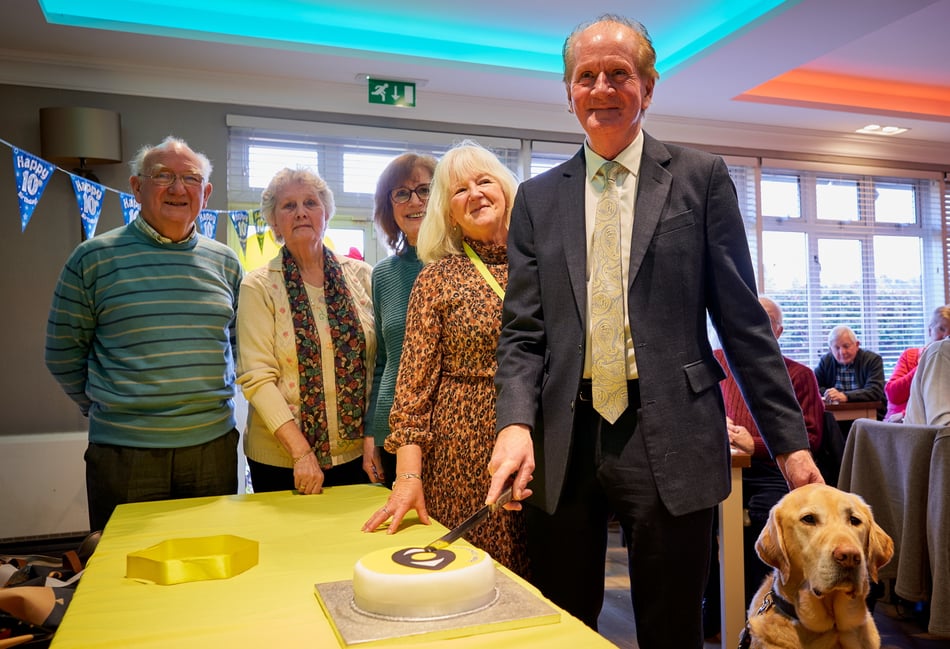 <p>Tom McNulty cut the cake with guide dog Toby and the volunteers who have been with the group for ten years</p>