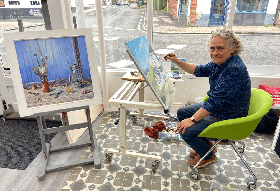 <p>Tim Burns works on his Aldeburgh painting next to one of his completed still life pictures</p>
